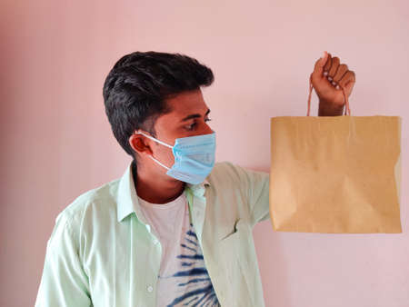 Young South Indian man wearing mask is seeing a shopping bag. Isolated on pink background. Shopping...