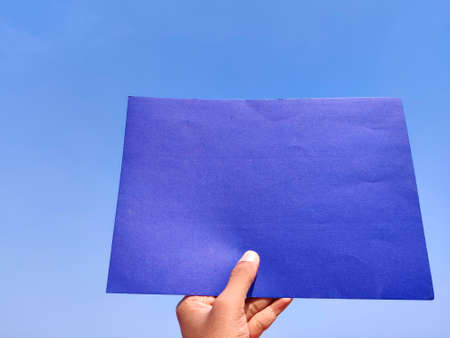 South Indian man hand holding empty dark blue sheet or paper. Cloud background.の写真素材