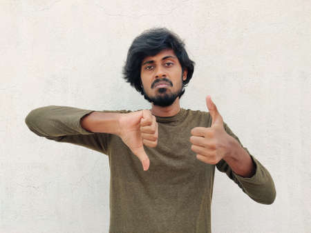 Young good looking man with green color full sleeves making good - bad sign. Thumbs up and thumbs down. White backgroundの写真素材