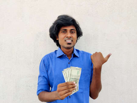 Clean shaved young handsome man wearing blue shirt holding four 500 rupees notes and pointing with thumb up to the side with happy face smiling. White backgroundの写真素材