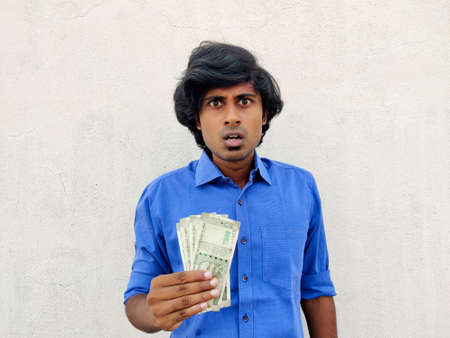 Shocking young handsome Indian man in blue shirt holding four 500 rupees notes. White backgroundの写真素材