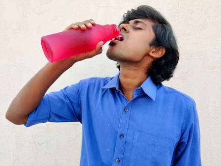 Closeup of young indian handsome man drinking water from pink bottle. Summer concept. White backgroundの写真素材