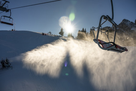 Snow cannon in alps, dolomites. Snow cannon or snow gun throwing snow on the ski slopes. The sun and mountains in the background.の写真素材
