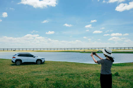 Asian woman take a picture and posed against white car in forest road with lake and bridge.の写真素材
