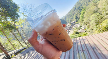 A hand holding a refreshing iced coffee in a scenic outdoor setting. The view showcases lush greenery and a tranquil village, perfect for enjoying a summer day.の写真素材