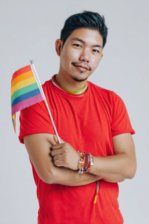 A young man proudly holds a rainbow flag in a studio setting, symbolizing diversity and inclusion. His joyful expression reflects the spirit of pride and support for the LGBTQ community.の素材