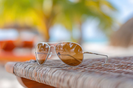 A pair of stylish sunglasses rests on a woven beach lounge, surrounded by tropical vibes. This image captures summer relaxation and sunny enjoyment, evoking a serene getaway atmosphere.の写真素材