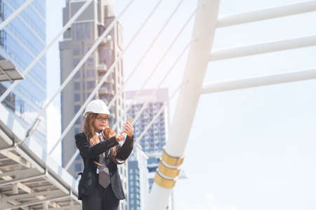 Middle-aged Engineer woman holding smart phone wearing construction hard hat or helmet. Middle-aged women professional standing in central of cityの写真素材