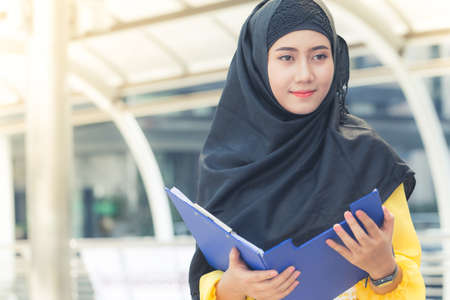 Young asian muslim business woman smiling and holding file document and standing at capital city. Beautiful muslim women. Thai muslim women.の写真素材
