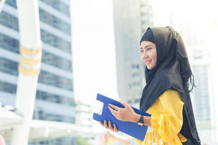 Young asian muslim business woman smiling and holding file document and standing at capital city. Beautiful muslim women. Thai muslim women.の写真素材