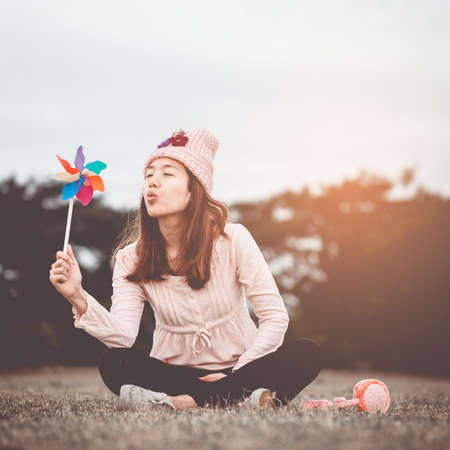 Beautiful girl holding wind toy or wind turbine or pinwheel and wool hat sitting on meadow in winter season in morning.の写真素材