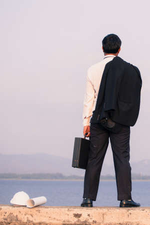 Back view of businessman in a suit with a briefcase and standing on concrete of reservoir with mountains view. Tired or stressed businessman concept.の写真素材