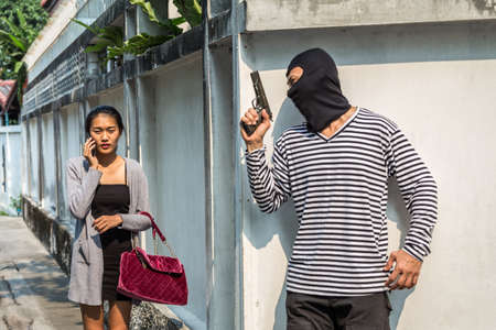 Man in hoodie robbing and threatening with gun to frightened young woman While use on the phone. criminal and robbery for woman concept.の写真素材