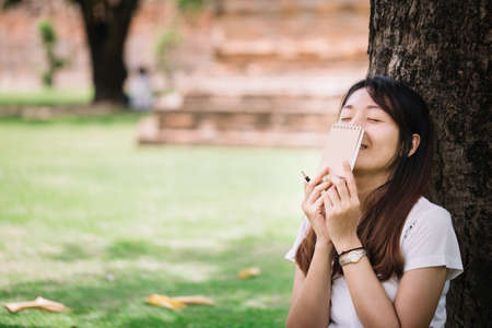young woman sitting under a tree and holding her diary with smile. women with natural conception.の写真素材