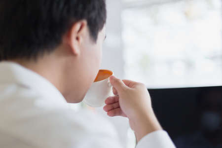 Young Asian businessman sipping fresh coffee and working by laptop at cafe in morning. Nomad working concept. Selective focus.の写真素材