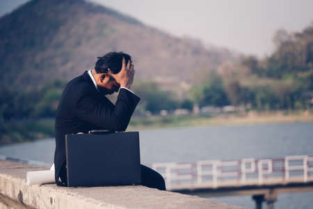 Back view of tired or stressed businessman in a suit with a briefcase and sitting on concrete of reservoir with mountains view. Unemployed businessman concept.の写真素材