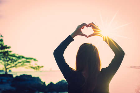 Silhouette of Heart shape being made by young woman hands with sunlight on beach. Sunset at the sea. Hand-made heart in love concept.の写真素材