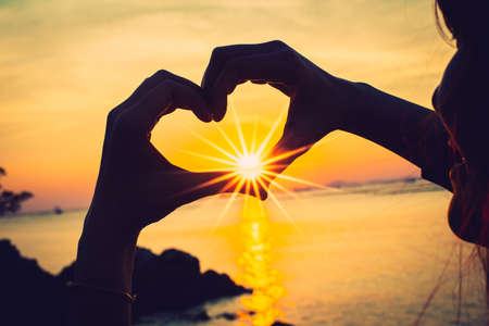 Silhouette of Heart shape being made by young woman hands with sunlight on beach. Sunset at the sea. Hand-made heart in love concept.の写真素材