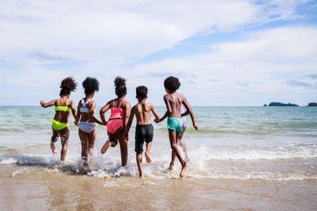 African American, Kids group in swimwear enjoying running to play the waves on beach. Ethnically diverse concept. Having fun after unlocking down from COVID 19. Summer holidays on beach with friendsの写真素材