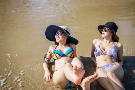 Outdoor summer portrait of the front image of two Asian beautiful girls in a sexy bikini lie down on the beach in summer. teenage girls best friends. sexy body. concept vacation, holiday in summerの写真素材