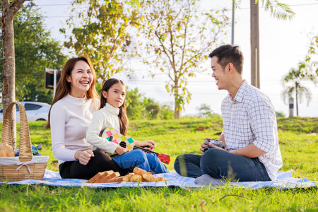 Happy family picnic. A little girl playing ukulele with her parents (Father, Mother) during picnicking on a picnic cloth on a sunny day. family relaxes in the green park. Family weekendの写真素材
