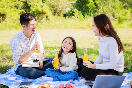 Happy family picnic. Asian parents (Father, Mother) and little girl drinking orange juice and have fun and enjoyed ourselves together during picnicking on a picnic cloth in the green gardenの写真素材
