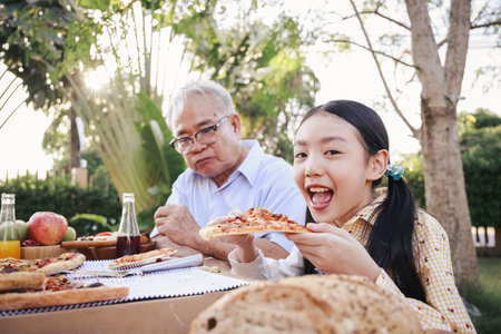 Asian retirement grandfather and pretty granddaughter enjoying to eating pizza together in home garden. Happy senior life after retirement with family concept.の写真素材