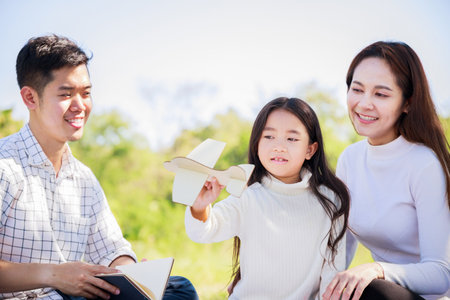 Happy family picnic. Asian parents (Father, Mother) and daughter playing the toy airplane and have enjoyed ourselves together while picnicking on picnic cloth in green garden in the sunshine dayの写真素材