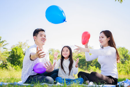 Happy family picnic. Asian parents (Father, Mother) and little girl playing balloon and have enjoyed ourselves together during picnicking on a picnic cloth in the green gardenの写真素材