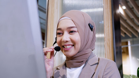 Beautiful woman in hijab at the computer with headset, distant work, call center. Confident islam woman worker in call center crossed arms face camera smiling joyful operator and helps with the order.の写真素材