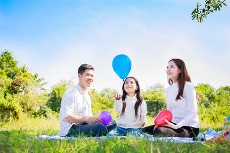 Happy family picnic. Asian parents (Father, Mother) and little girl playing balloon and have enjoyed ourselves together during picnicking on a picnic cloth in the green gardenの写真素材