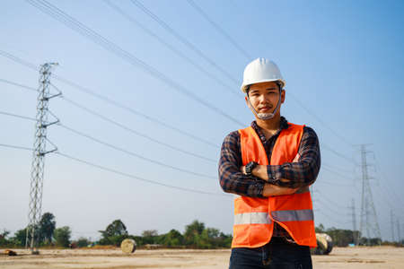 Portrait of Electrical Engineer, field engineer, foreman, owner standing in construction site project and Electric power line and pole in background. Asian engineer. High voltage power line pylon.の写真素材