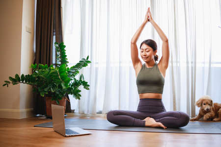 Asian woman practicing yoga from yoga online course on a video conference on a laptop at home. Healthy lifestyle - technology at home. New normal lifestyleの写真素材