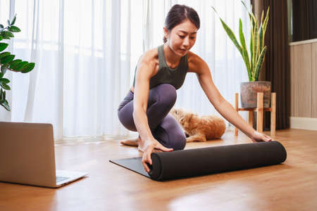 young Asian woman unrolling roll black yoga mat for online training yoga with her dog at home by laptop for quarantine herself from Covid-19, New normal lifestyle conceptの写真素材