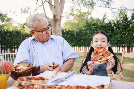 Asian retirement grandfather and pretty granddaughter enjoying to eating pizza together in home garden. Happy senior life after retirement with family concept.の写真素材