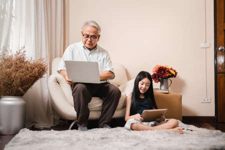Asian retirement grandfather using a laptop and sitting on sofa while pretty granddaughter doing homework in tablet in living room. Family educational at home concept. Technology and education.の写真素材