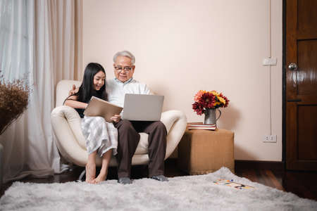Asian retirement grandfather teaching homework pretty granddaughter by tablet and laptop in the living room. Happy Family educational at home concept. Technology and education.の写真素材