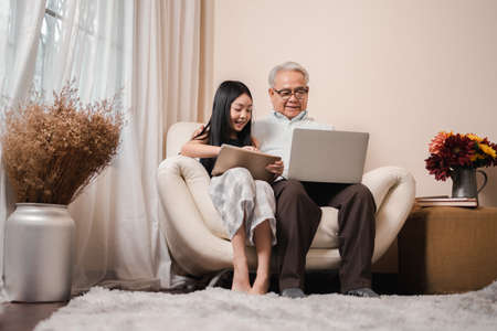 Asian retirement grandfather teaching homework pretty granddaughter by tablet and laptop in the living room. Happy Family educational at home concept. Technology and education.の写真素材