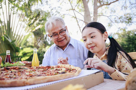 Asian retirement grandfather and pretty granddaughter enjoying to eating pizza together in home garden. Happy senior life after retirement with family concept.の写真素材