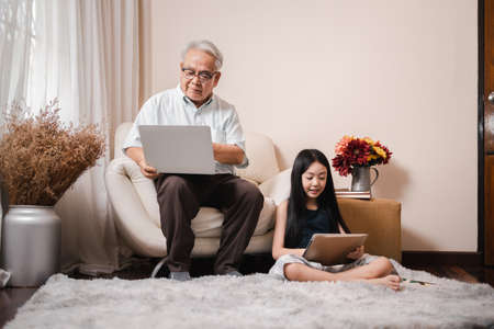 Asian retirement grandfather using a laptop and sitting on sofa while pretty granddaughter doing homework in tablet in living room. Family educational at home concept. Technology and education.の写真素材