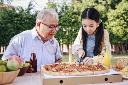 Granddaughter enjoying slicing pizza for retirement grandfather in home garden. Happy senior life after retirement with family concept.の写真素材