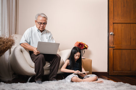 Asian retirement grandfather using a laptop and sitting on sofa while pretty granddaughter doing homework in tablet in living room. Family educational at home concept. Technology and education.の写真素材