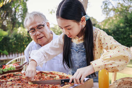 Granddaughter enjoying slicing pizza for retirement grandfather in home garden. Happy senior life after retirement with family concept.の写真素材