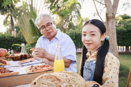 Asian retirement grandfather and pretty granddaughter enjoying to eating pizza together in home garden. Happy senior life after retirement with family concept.の写真素材