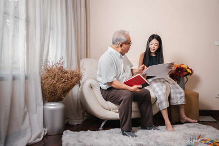 Asian retirement grandfather teaching and checking drawing on a paper with pretty granddaughter together while holding book and sits on the sofa in living room. Family education at home. Home school.の写真素材
