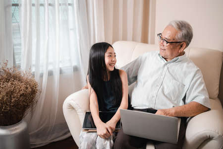 Happy Asian retirement grandfather and pretty granddaughter laughing while watching e-book for learning to education together by laptop. Family educational at home concept. Technology and education.の写真素材