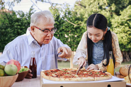 Granddaughter enjoying slicing pizza for retirement grandfather in home garden. Happy senior life after retirement with family concept.の写真素材