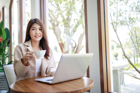attractive Asian businesswoman drinking coffee and working by laptop in the coffee shop. Confident and smart woman and business concept.の写真素材