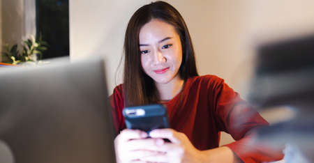 Happy female freelancer using smartphone during overtime at home office. Busy people working.の写真素材