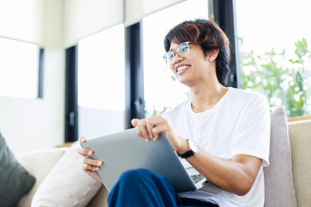 Smiling man sitting on couch at home using laptop for remote working from home office in living roomの写真素材